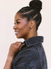 A woman in profile wearing a dark denim jacket and a small silver stud earring, with her hair styled in a high bun and cornrow braids, against a solid light beige background.