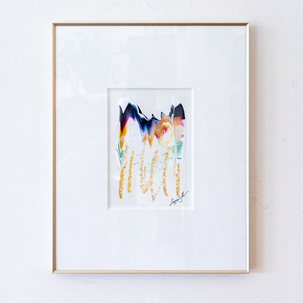 A framed abstract art print featuring colorful vertical brushstrokes in gold, pink, blue, and black on a white mat, enclosed in a thin gold frame and hung on a white textured wall.