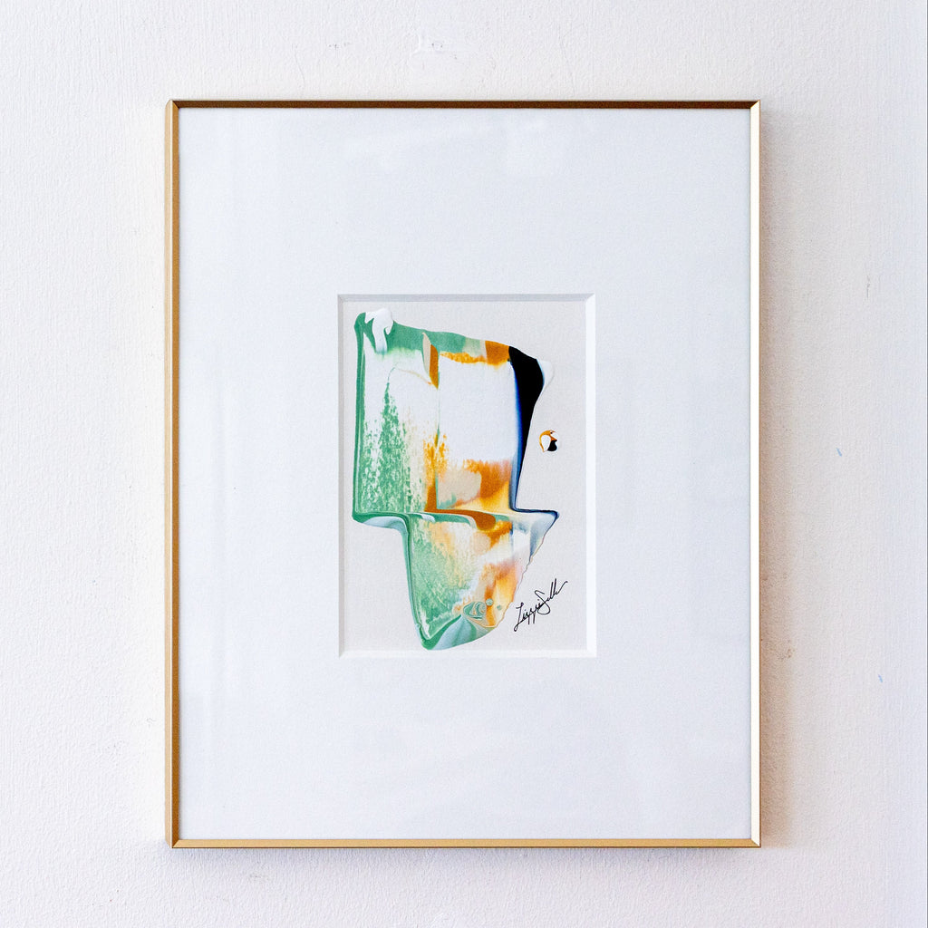 A framed abstract art print featuring green, orange, and black paint strokes on a white mat, enclosed in a thin gold frame against a white wall.