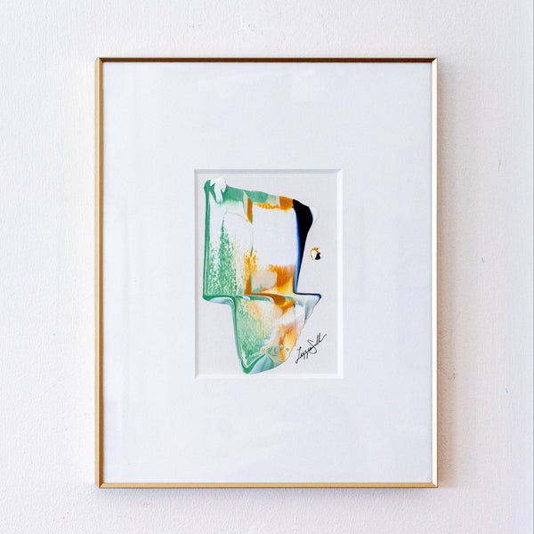 A framed abstract art print featuring green, orange, and black paint strokes on a white mat, enclosed in a thin gold frame against a white wall.