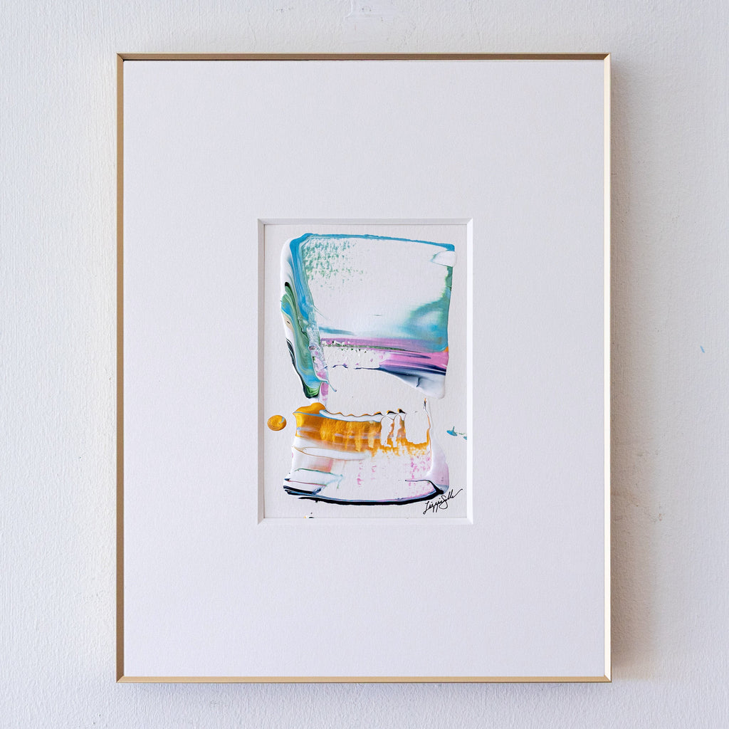 A framed abstract painting, set in a white mat and a thin gold frame against a white wall.