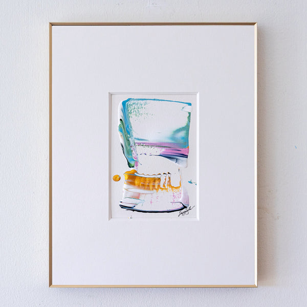 A framed abstract painting, set in a white mat and a thin gold frame against a white wall.