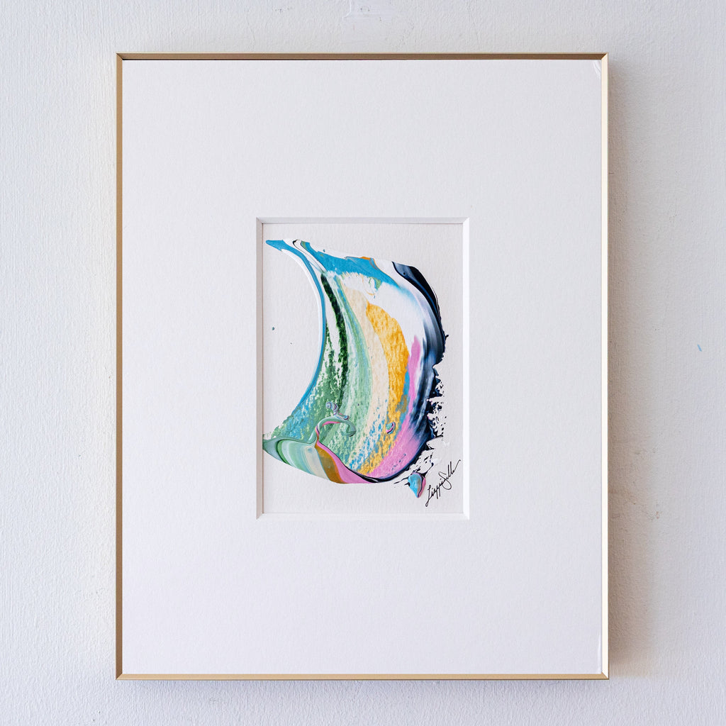A framed abstract artwork featuring a colorful fluid swirl design in shades of blue, green, yellow, orange, pink, and black, presented in a white mat and a thin gold frame against a white wall.