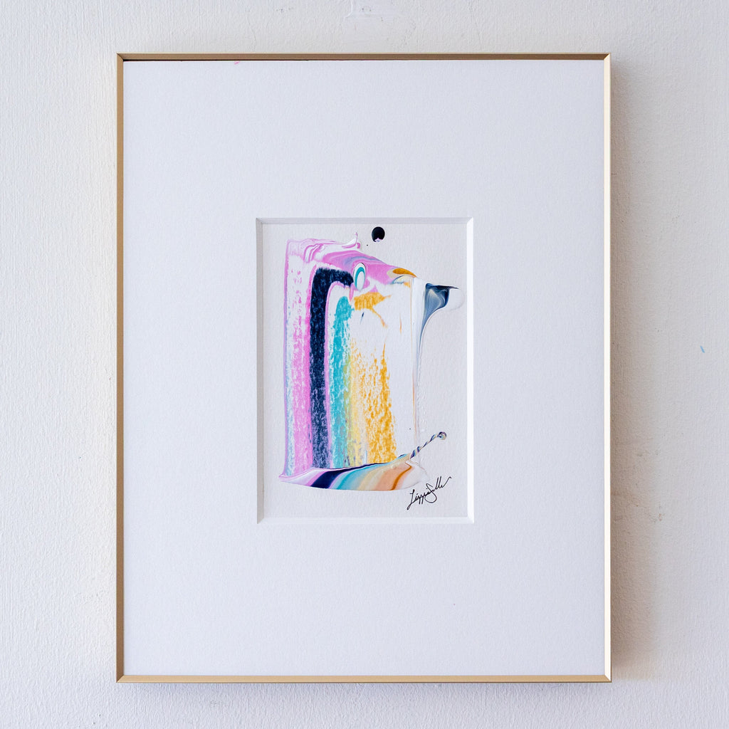 A framed abstract artwork, set within a white mat and a thin gold frame on a light gray wall.