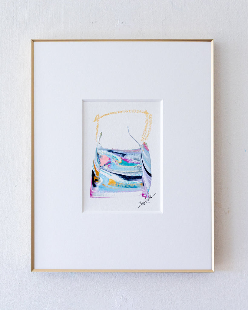 A framed abstract artwork, set in a white mat and a thin gold frame against a light textured wall.