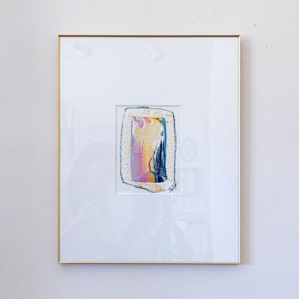 A framed abstract painting featuring a colorful, textured vertical rectangle in shades of pink, yellow, blue, and purple, set within a wide white mat and a thin gold frame, mounted on a light gray textured wall.