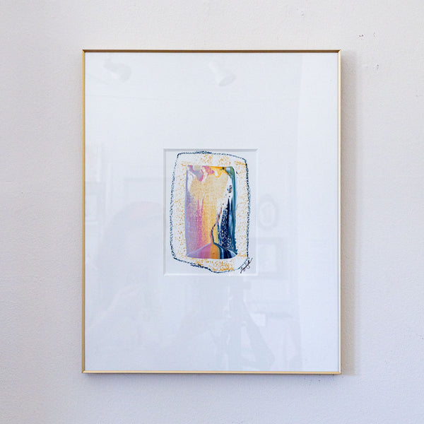 A framed abstract painting featuring a colorful, textured vertical rectangle in shades of pink, yellow, blue, and purple, set within a wide white mat and a thin gold frame, mounted on a light gray textured wall.