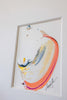 An abstract framed painting featuring a curved, organic shape in orange, yellow, blue, and white, mounted on a light gray wall.