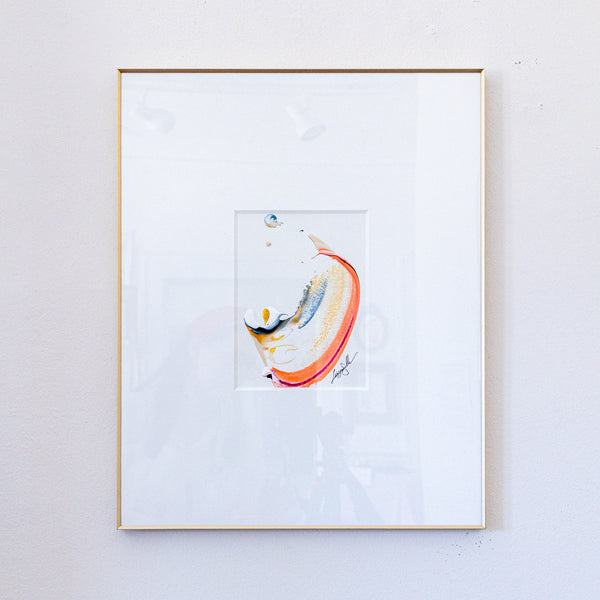 A framed abstract painting featuring a minimalist splash of orange, yellow, blue, and white paint on a white mat, enclosed in a thin gold frame and hung on a light gray wall.