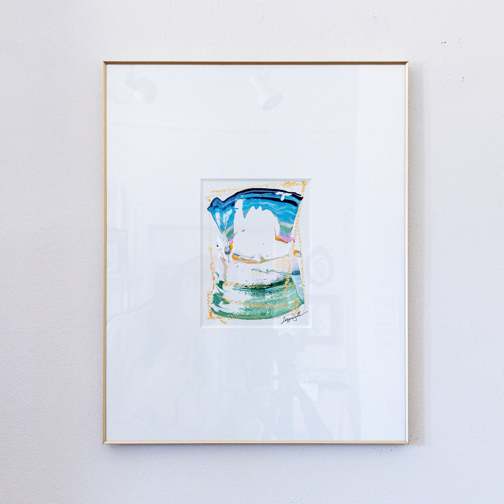 A framed painting featuring a colorful acrylic painting with blue, green, and white tones, set in a white mat and a thin gold frame against a white wall.