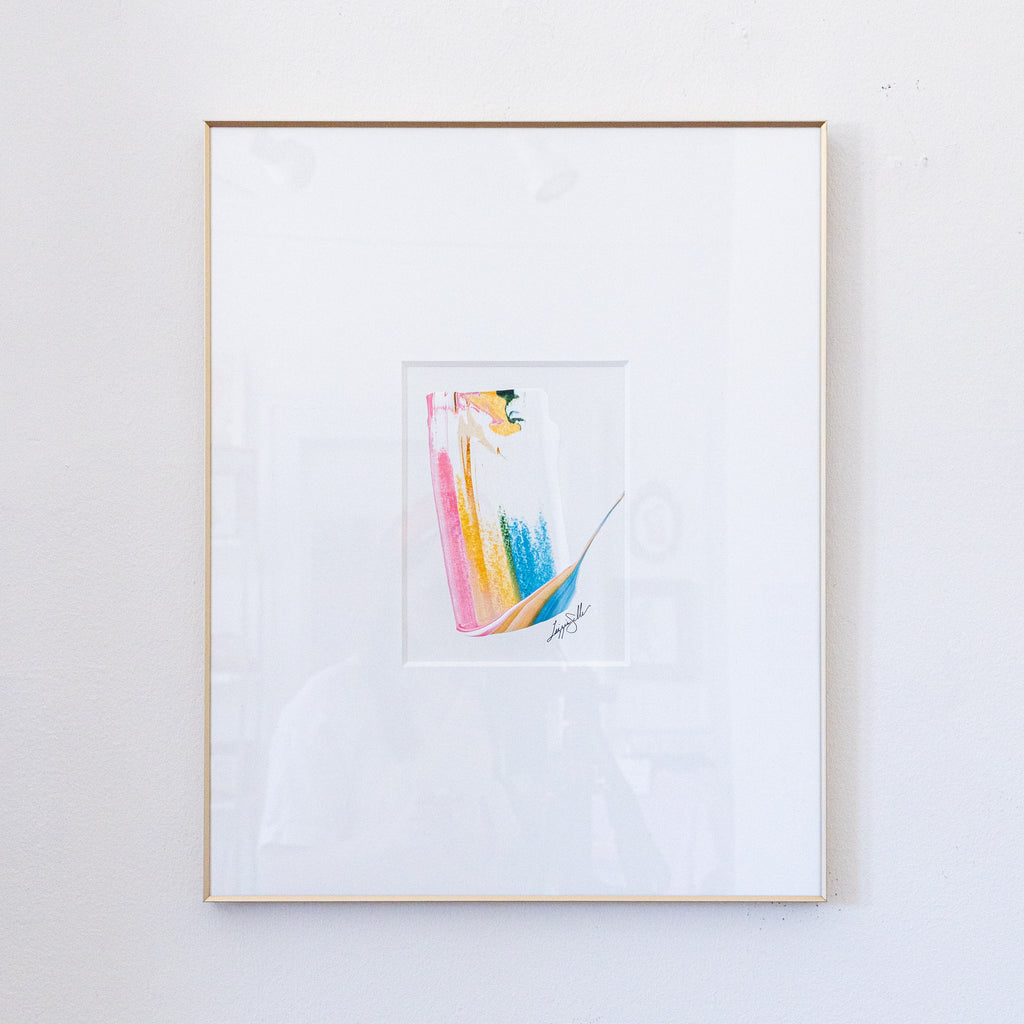 A framed abstract painting featuring a minimalist colorful figure in pink, gold, blue, and green on a white mat, enclosed in a light wood frame against a white wall.