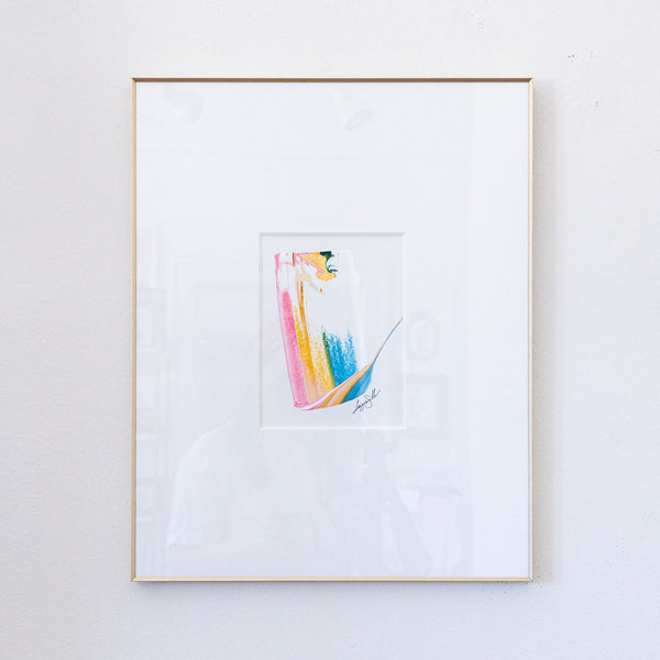 A framed abstract painting featuring a minimalist colorful figure in pink, gold, blue, and green on a white mat, enclosed in a light wood frame against a white wall.