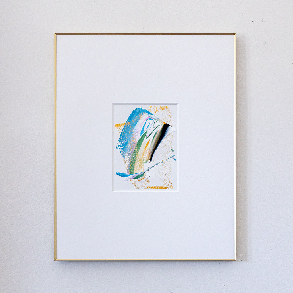 A framed abstract painting featuring blue, green, black, and gold in a white mat, enclosed in a thin gold frame and hung on a light gray wall.