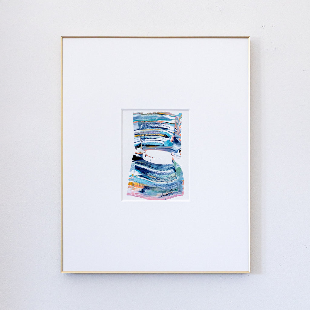 A framed abstract painting featuring textured blue, green, and pink brushstrokes, presented in a white mat and a thin gold frame against a light gray wall.