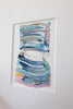 An abstract framed painting featuring colorful painted brushstrokes in shades of blue, green, pink, yellow, and white, mounted on a plain white wall.