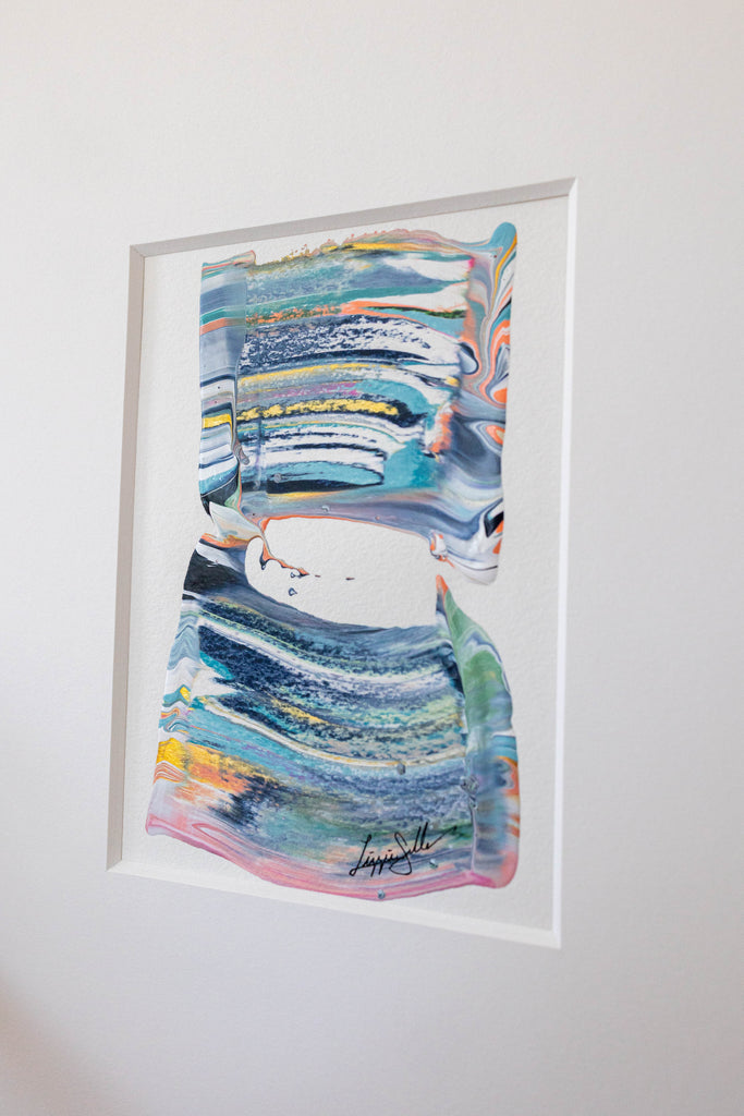 An abstract framed painting featuring colorful painted brushstrokes in shades of blue, green, pink, yellow, and white, mounted on a plain white wall.
