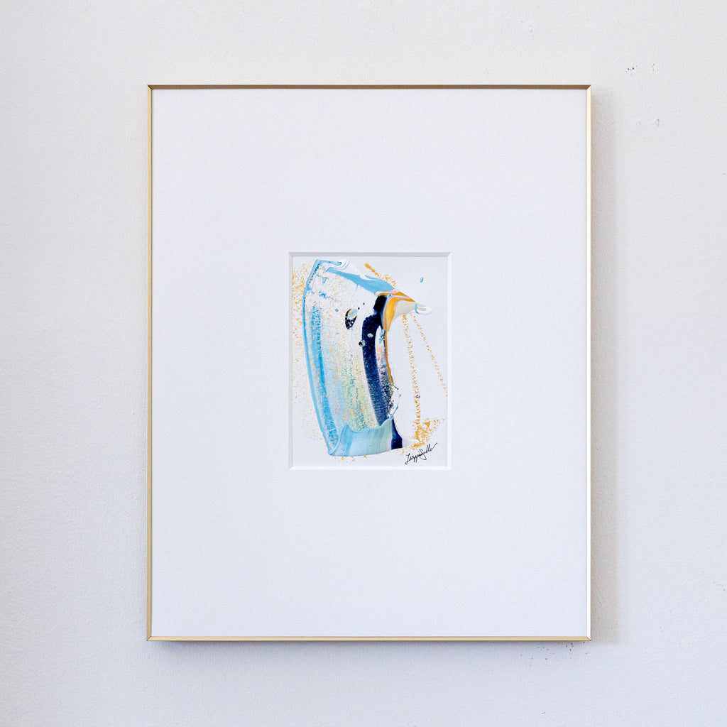 A framed abstract painting featuring a vertical swish in shades of blue, white, and yellow, presented in a white mat and a thin gold frame against a light gray wall.