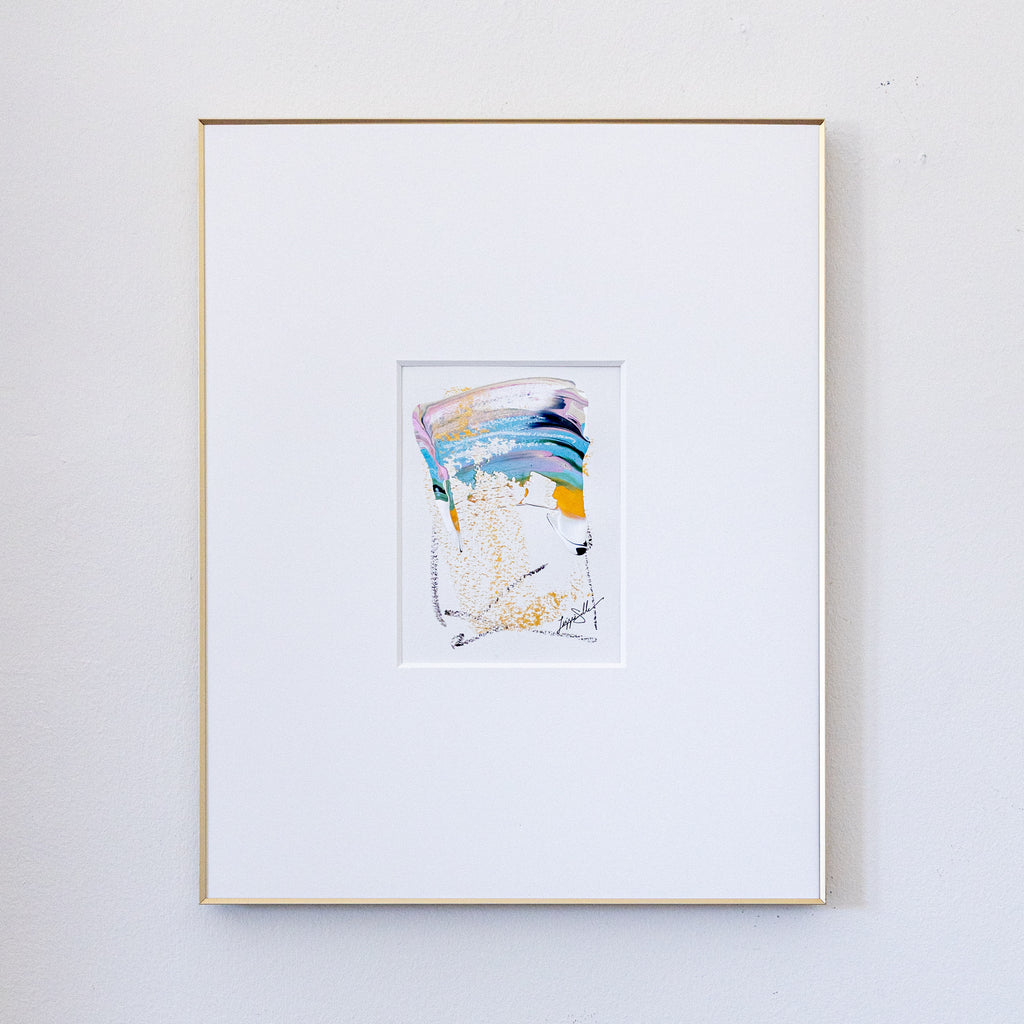 A framed abstract painting featuring blue, metallic gold, light pink, and black oil pastel markings, presented in a white mat and a thin gold frame against a light gray wall.
