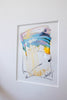 A framed abstract painting featuring blue, metallic gold, light pink, and black oil pastel markings, presented in a white mat.