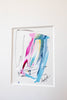A framed abstract painting featuring vibrant blue, pink, and green paint strokes on a white background, hung on a light gray wall.