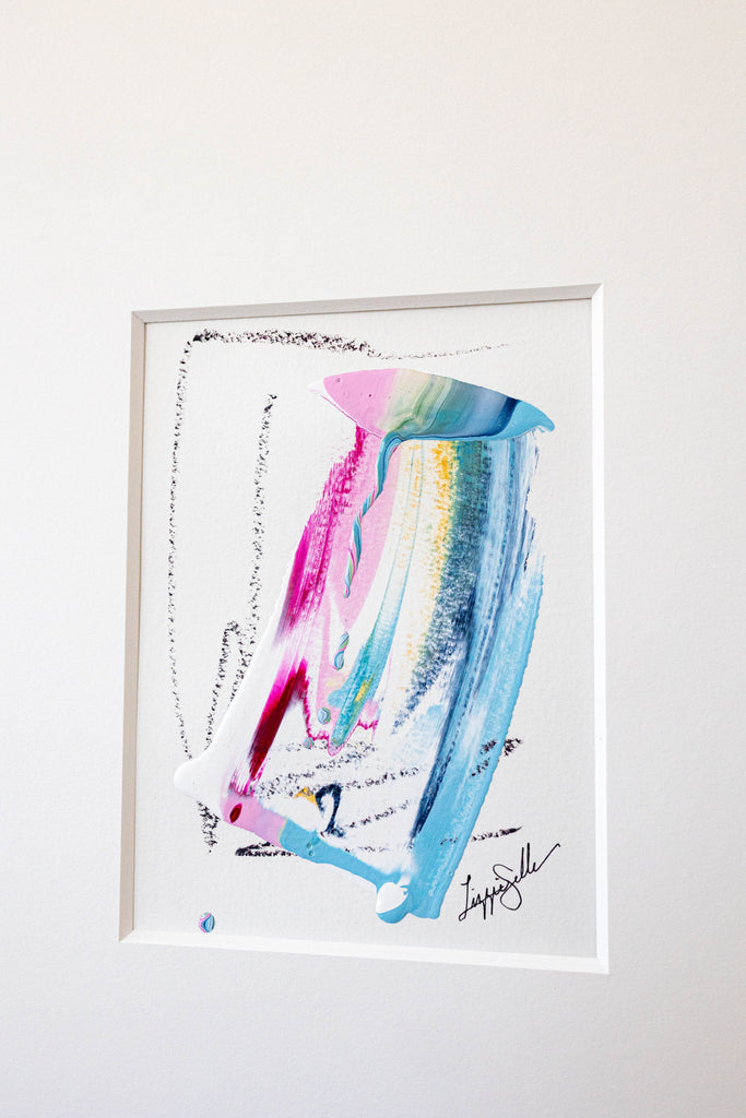 A framed abstract painting featuring vibrant blue, pink, and green paint strokes on a white background, hung on a light gray wall.