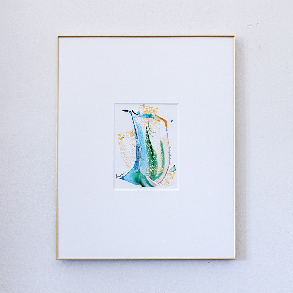 A framed painting featuring a central colorful composition of blue, green, and gold brushstrokes on a white mat, enclosed in a thin gold frame against a light gray wall.