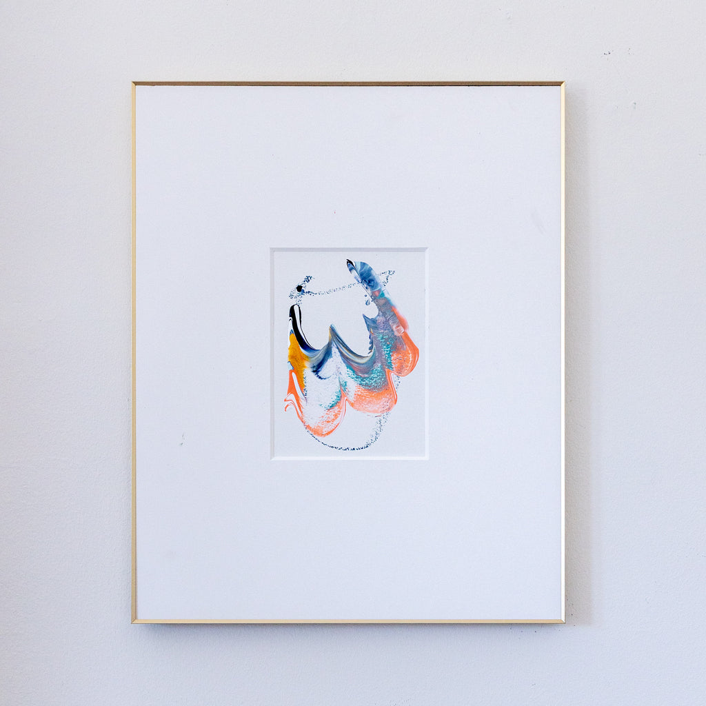 A framed abstract painting featuring blue, coral, blue, gold, and black in a white mat, enclosed in a thin gold frame against a light gray wall.