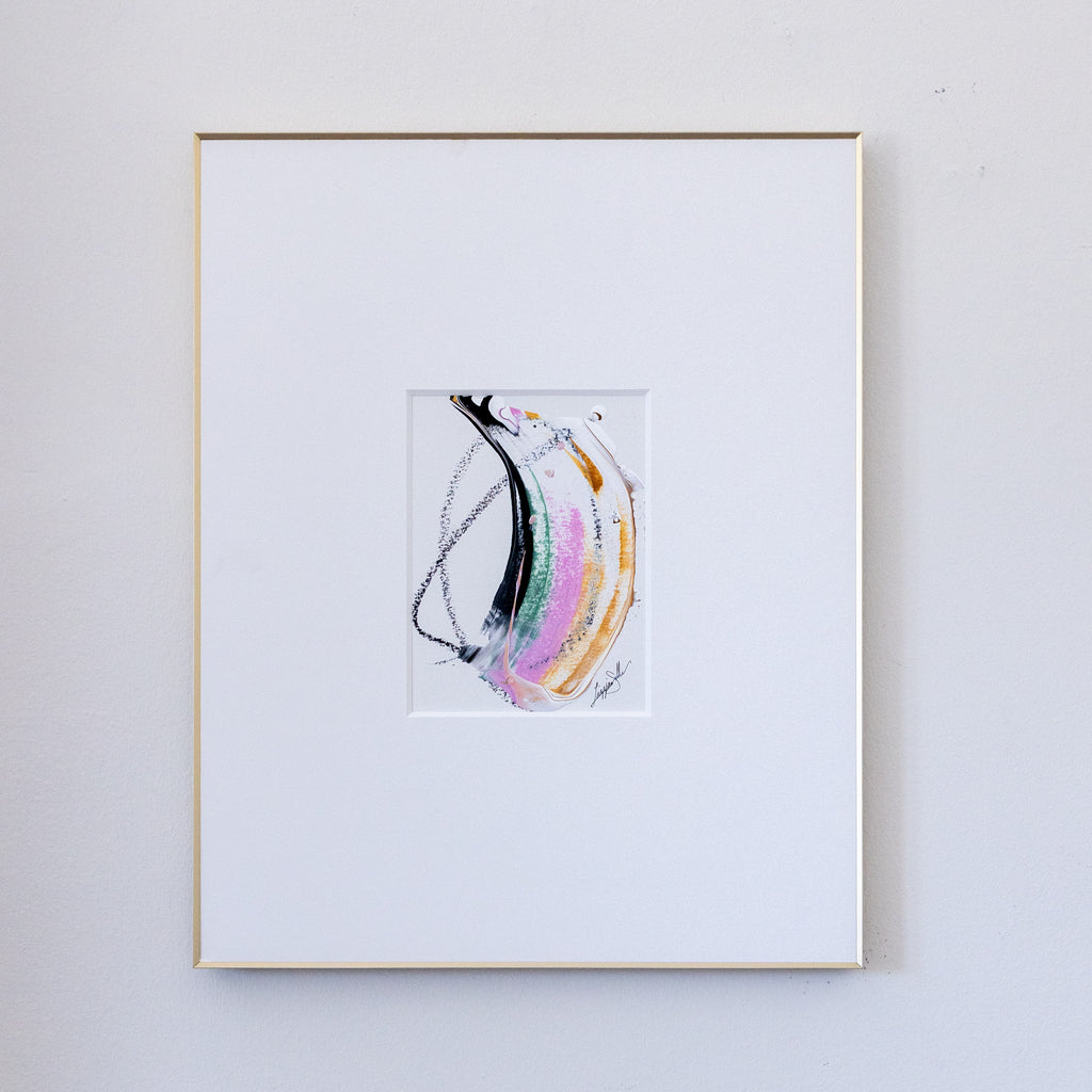 A framed abstract painting featuring fluid, acrylic in black, pink, metallic gold and green with black oil pastel markings in a white mat, enclosed in a thin gold frame against a light gray wall.