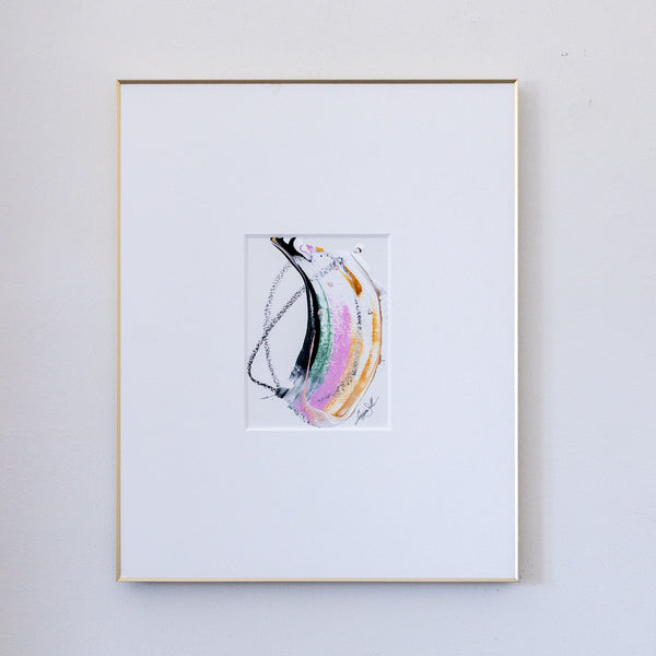A framed abstract painting featuring fluid, acrylic in black, pink, metallic gold and green with black oil pastel markings in a white mat, enclosed in a thin gold frame against a light gray wall.
