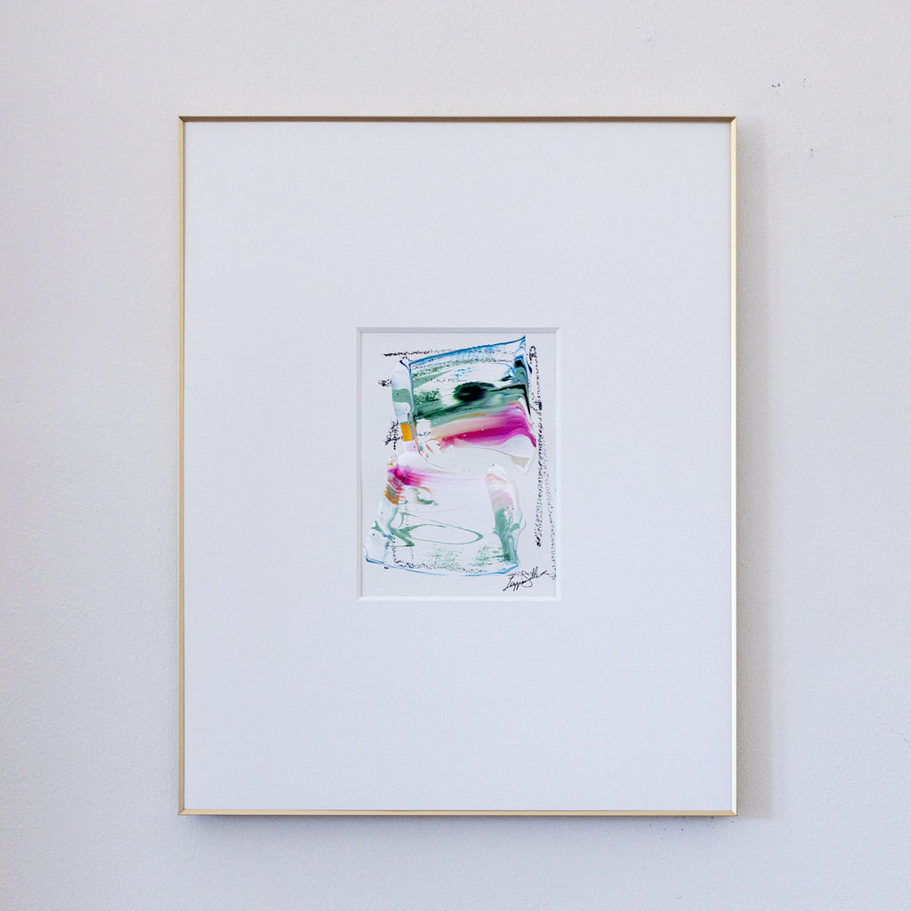 A framed abstract painting featuring colorful acrylic brushstrokes, presented in a white mat and a thin gold frame against a light gray wall.