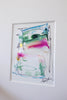 An abstract framed painting featuring fluid, layered paint textures in green, pink, blue, and white, mounted on a plain white wall.