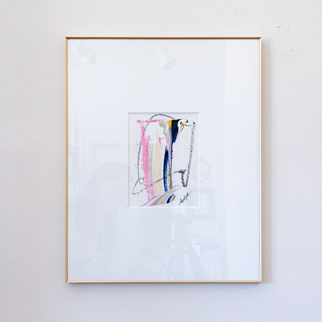 A framed abstract painting featuring pastel pink, yellow, blue, and black brushstrokes on a white mat, enclosed in a thin gold frame and hung on a light gray wall.