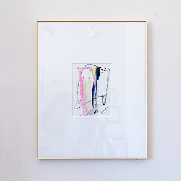 A framed abstract painting featuring pastel pink, yellow, blue, and black brushstrokes on a white mat, enclosed in a thin gold frame and hung on a light gray wall.