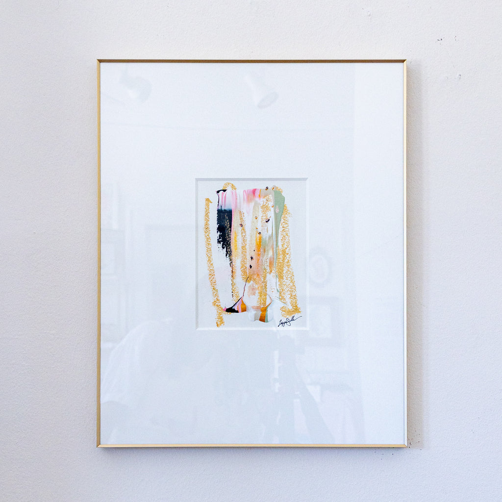 A framed abstract PAINTING featuring vertical brushstrokes in gold, pink, black, and green on a white mat, enclosed in a thin gold frame and hung on a light gray wall.