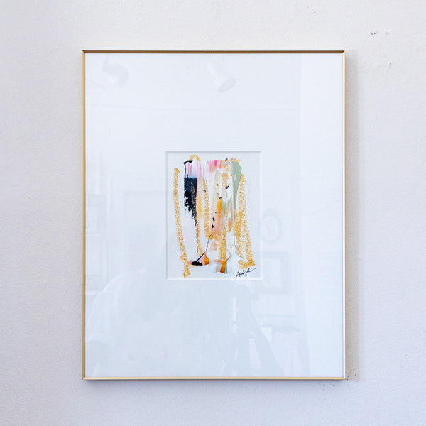 A framed abstract painting featuring vertical brushstrokes in gold, pink, black, and green on a white mat, enclosed in a thin gold frame and hung on a light gray wall.