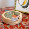 A round wooden coastertainer holding stacked coasters with a colorful floral and bird illustration, placed on a red floral tabletop with a decorative framed print in the background.