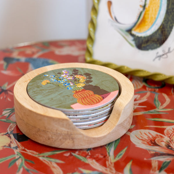 A round wooden coastertainer holding stacked coasters with a colorful floral and bird illustration, placed on a red floral tabletop with a decorative framed print in the background.
