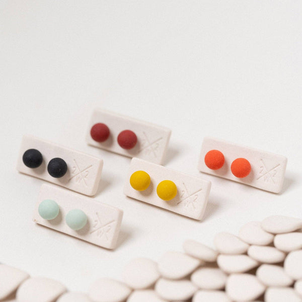 Five pairs of stud earrings displayed on small rectangular holders, each pair featuring two matte round studs in different colors: black, red, yellow, orange, and mint green. The holders are arranged on a white surface with a shallow depth of field focusing on the center pair, with a blurred row of white oval stones in the foreground.