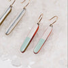 Three dangle earrings with elongated rectangular charms in different color combinations, displayed on a bed of white sea salt. The earrings feature gold-tone fishhook wires and charms with a silver-and-white split design, a red-and-white split design, and a mint-green-and-white split design.