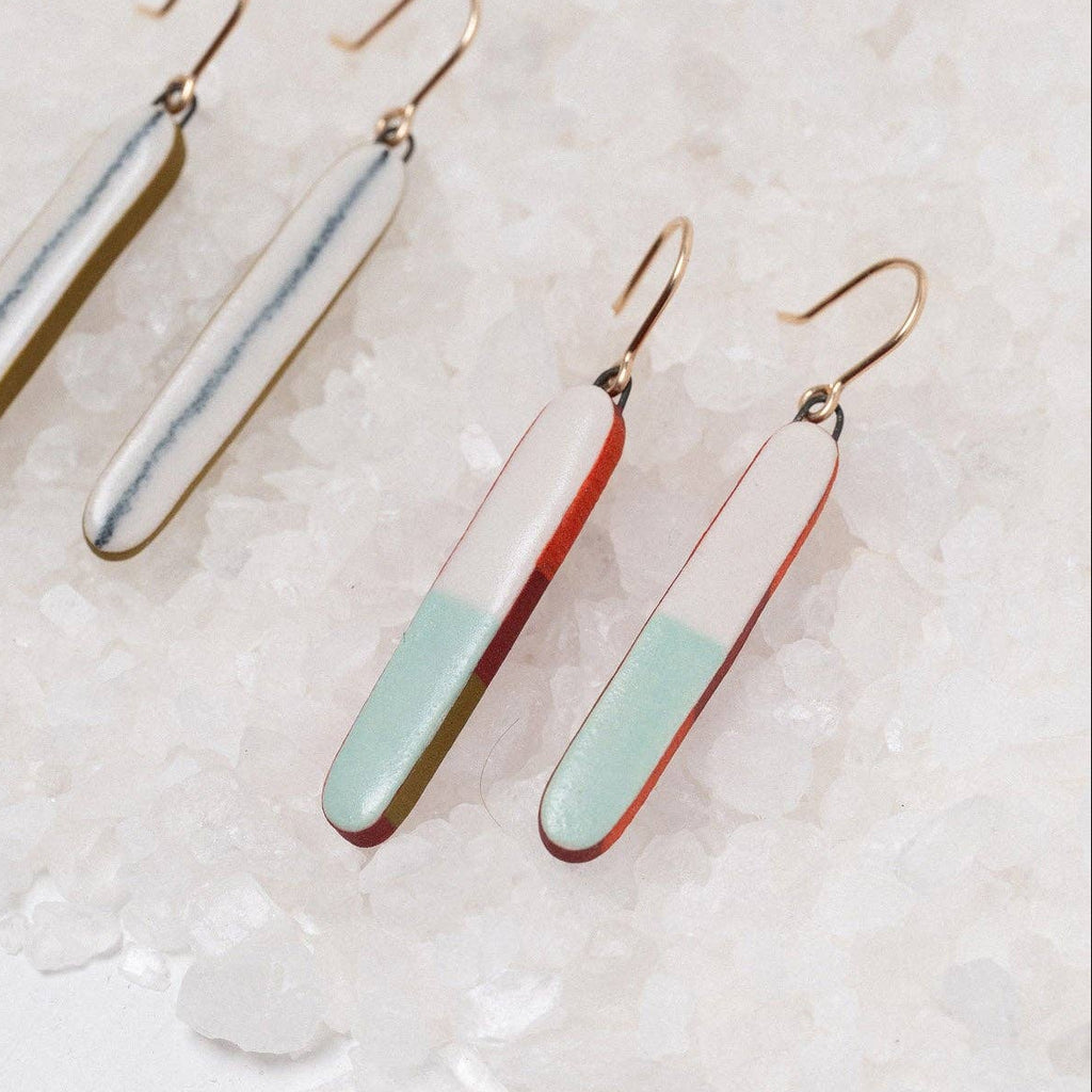 Three dangle earrings with elongated rectangular charms in different color combinations, displayed on a bed of white sea salt. The earrings feature gold-tone fishhook wires and charms with a silver-and-white split design, a red-and-white split design, and a mint-green-and-white split design.
