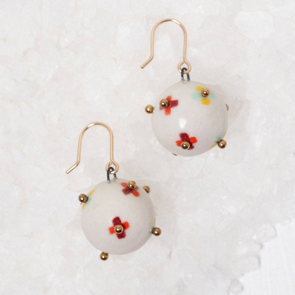 A pair of white spherical dangle earrings with small red and yellow floral details, gold-tone hooks, and tiny gold accent beads, displayed on a textured white surface.