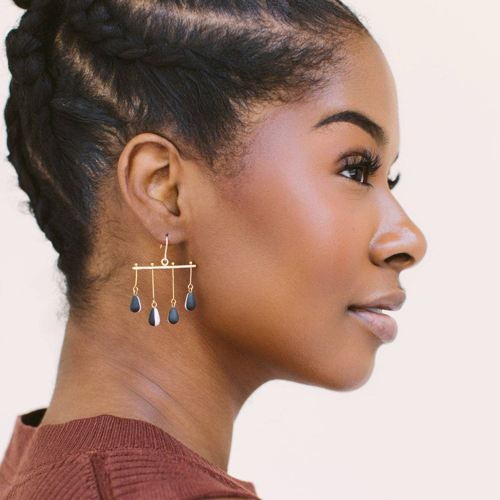 A close-up side profile of a woman wearing a pair of gold geometric dangle earrings with dark blue-gray teardrop charms, paired with a rust-colored ribbed top against a light beige background.