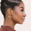 A close-up side profile of a woman wearing a pair of gold geometric dangle earrings with dark blue-gray teardrop charms, paired with a rust-colored ribbed top against a light beige background.