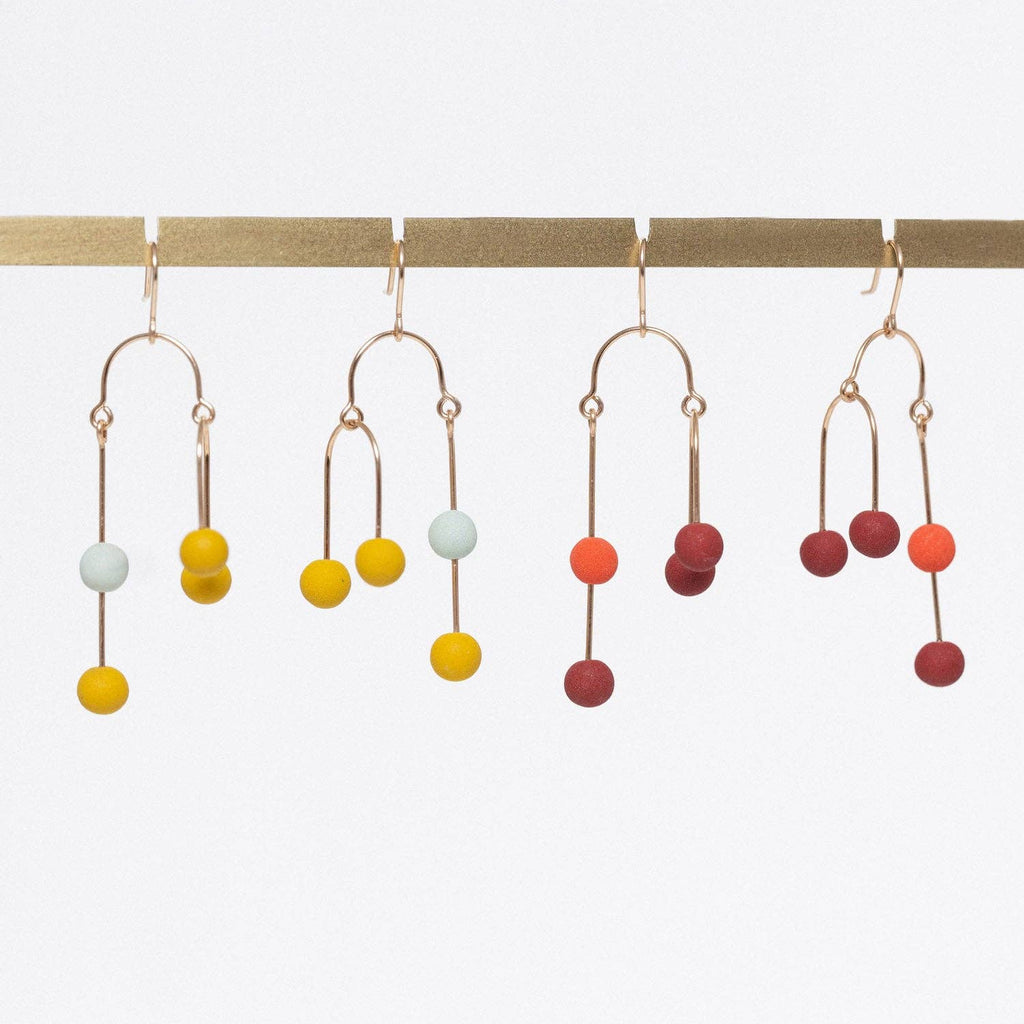 Four pairs of gold-tone dangle earrings with small matte-colored beads, hanging from a gold display bar against a white background.