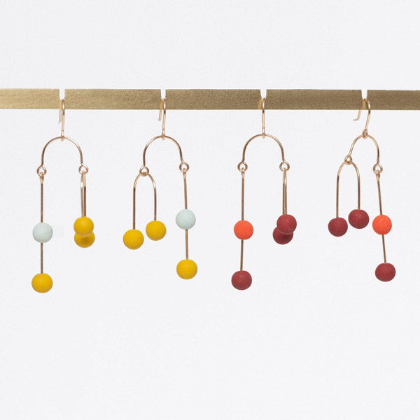 Four pairs of gold-tone dangle earrings with small matte-colored beads, hanging from a gold display bar against a white background.