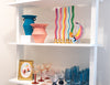 A white shelving unit displaying a collection of colorful glassware and decorative art. The shelves feature blue and pink ceramic vases, a rainbow-striped glass candle holder, a colorful glass bird figurine, and multiple styles of blue, red, and clear stemmed glasses.