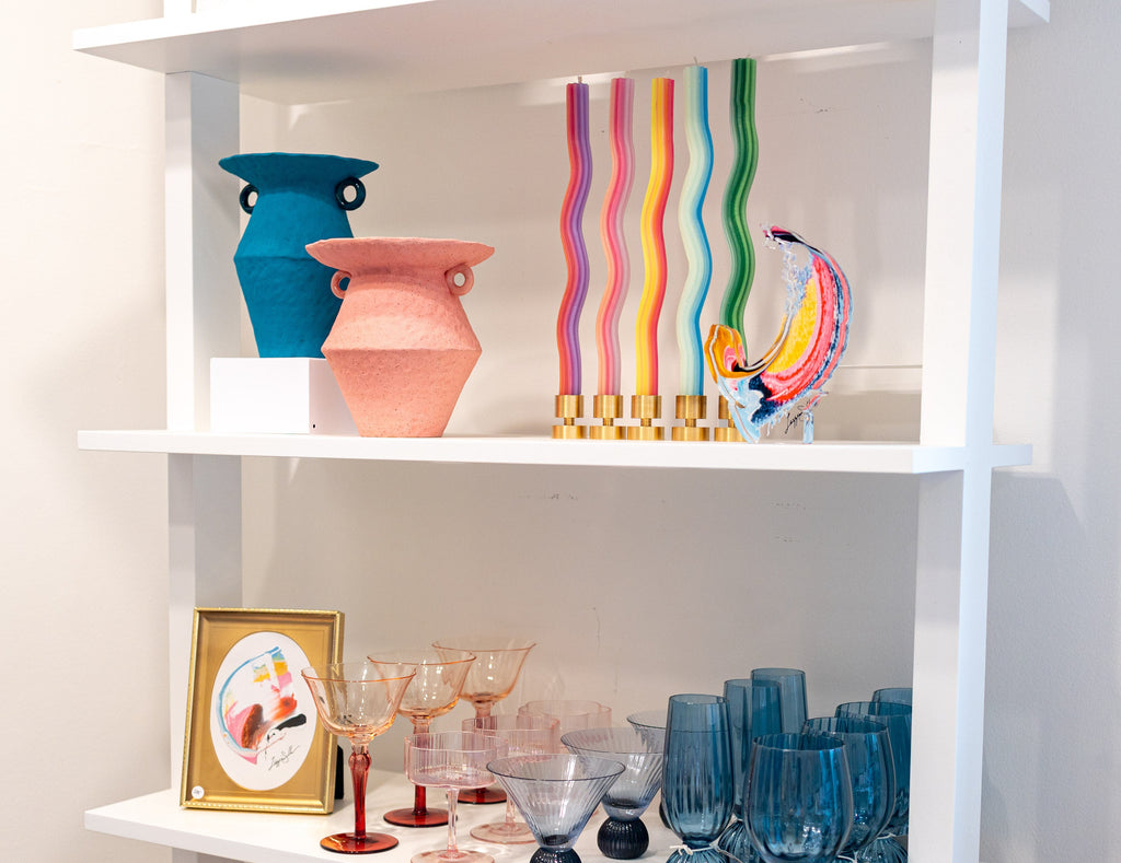 A white shelving unit displaying a collection of colorful glassware and decorative art. The shelves feature blue and pink ceramic vases, a rainbow-striped glass candle holder, a colorful glass bird figurine, and multiple styles of blue, red, and clear stemmed glasses.