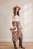 A woman stands in a studio wearing a beige wide-brim hat, a cream ribbed long-sleeve top, a brown and beige patterned midi skirt, and brown snakeskin boots. She carries a cream crossbody bag with a fringed handle.