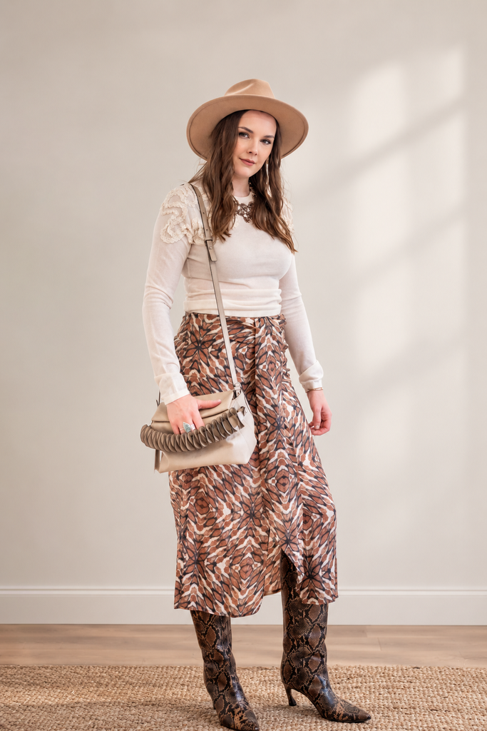 A woman stands in a studio wearing a beige wide-brim hat, a cream ribbed long-sleeve top, a brown and beige patterned midi skirt, and brown snakeskin boots. She carries a cream crossbody bag with a fringed handle.