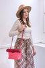 A woman wearing a beige wide-brim hat, a cream lace-trim long-sleeve top, and a brown patterned skirt carries a bright pink textured shoulder bag with a thin strap in a minimalist white room.
